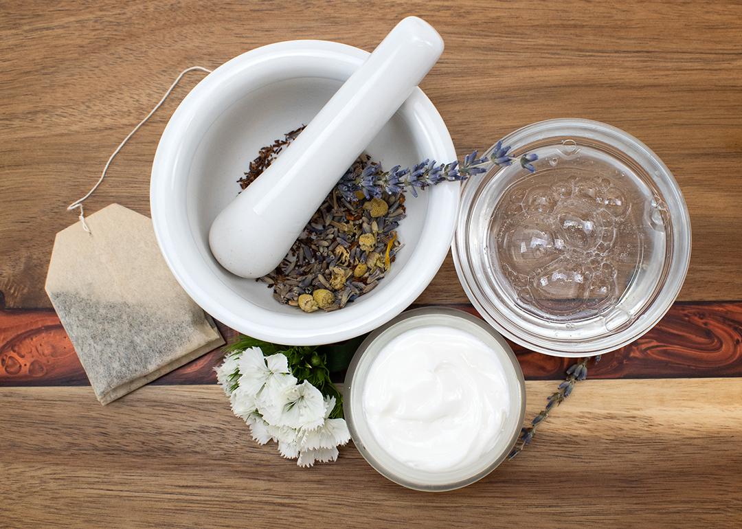 A top view of natural beauty ingredients on a table such as a tea bag, face cream and soap, and a mortar and pestle with dried flowers.