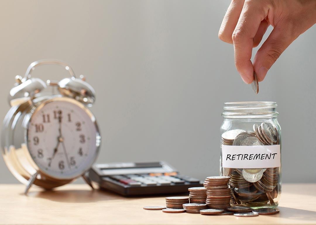Hand putting coins in glass jar labeled "retirement" next to calculator and analog alarm clock with window in above in the background. 