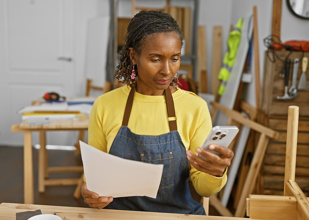 A black female carpentry business owner holds a document and reads information from her smartphone.