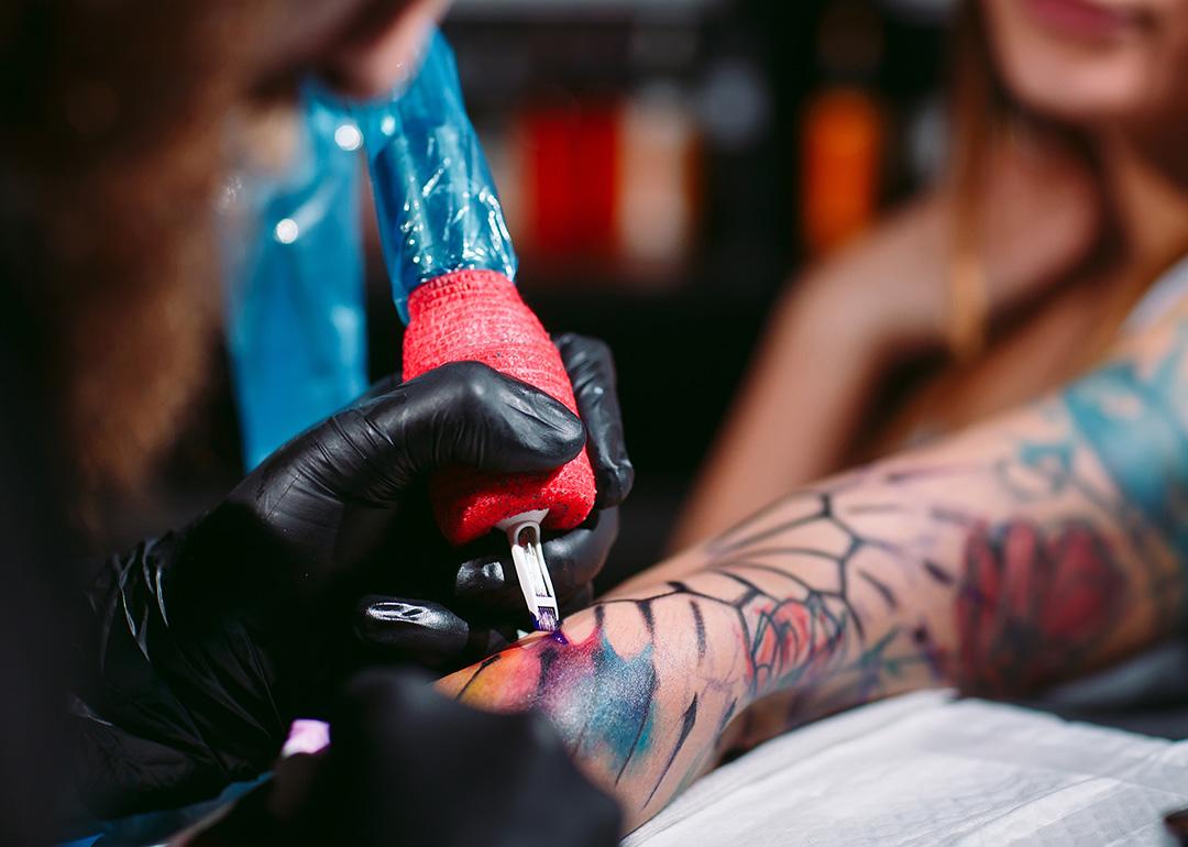A professional artist tattooing a design on a woman's left forearm.