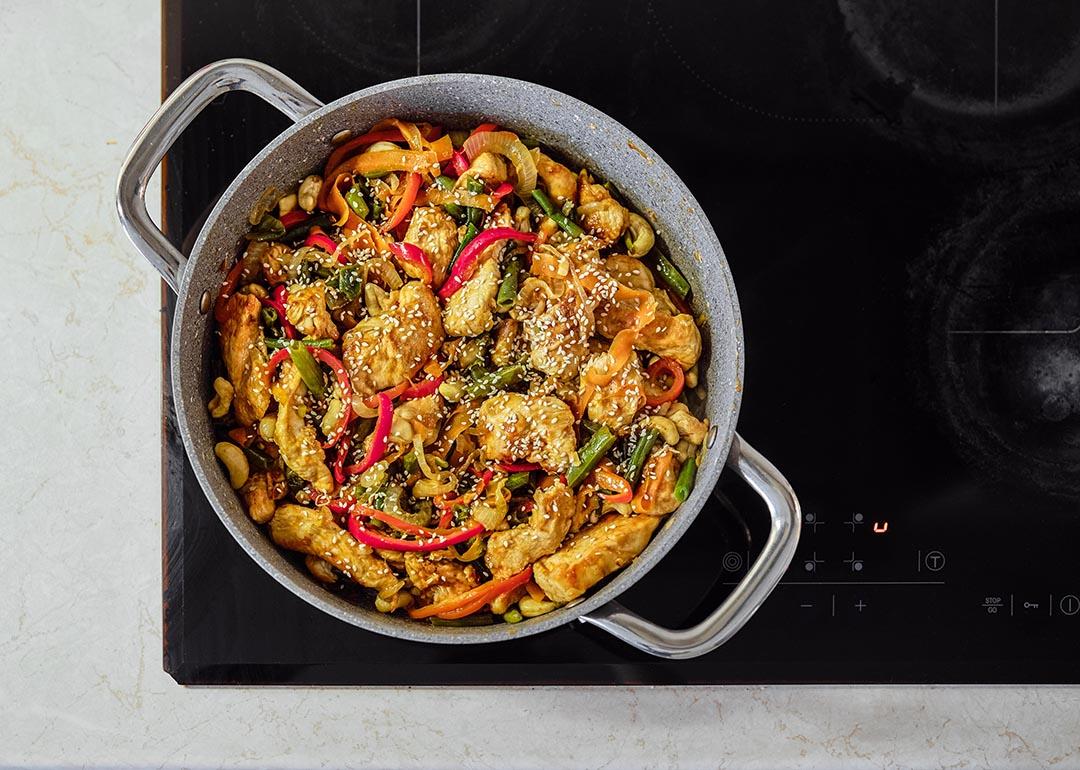 A colorful mix of stir-fried chicken, fresh vegetables, and cashews is sizzling in a pot on an induction cooktop.