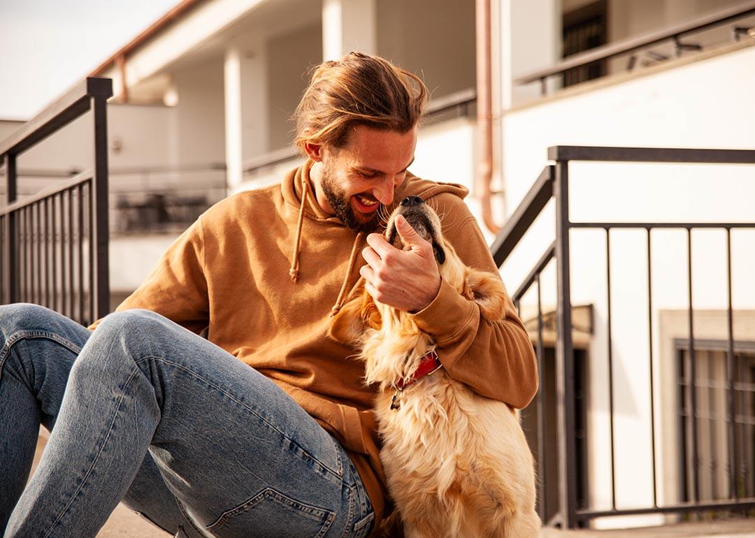 Sporty guy in honey-colored pullover hoodie and jeans sitting outside on steps playfully hugging a golden retriever.