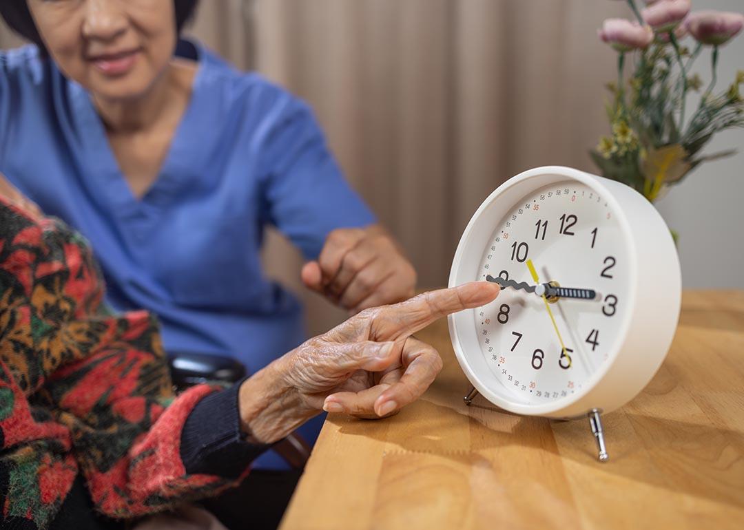 Close up of Asian elderly woman's hand pointing at analog clock with caregiver visible behind her.