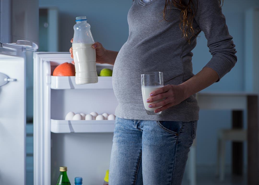 Pregnant woman, whose face is not shown, is illuminated by light of open fridge holding a glass in one hand and bottle of milk in the other.