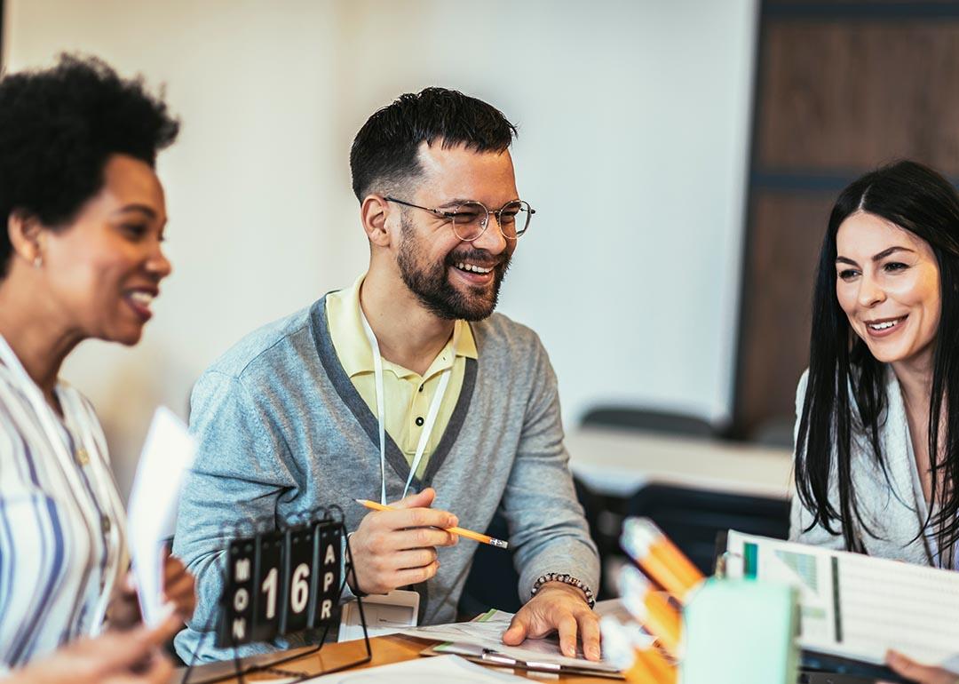 Group of three sitting at a table at work. Man in the middle is laughing and holding a pencil and two women are on either side of him.
