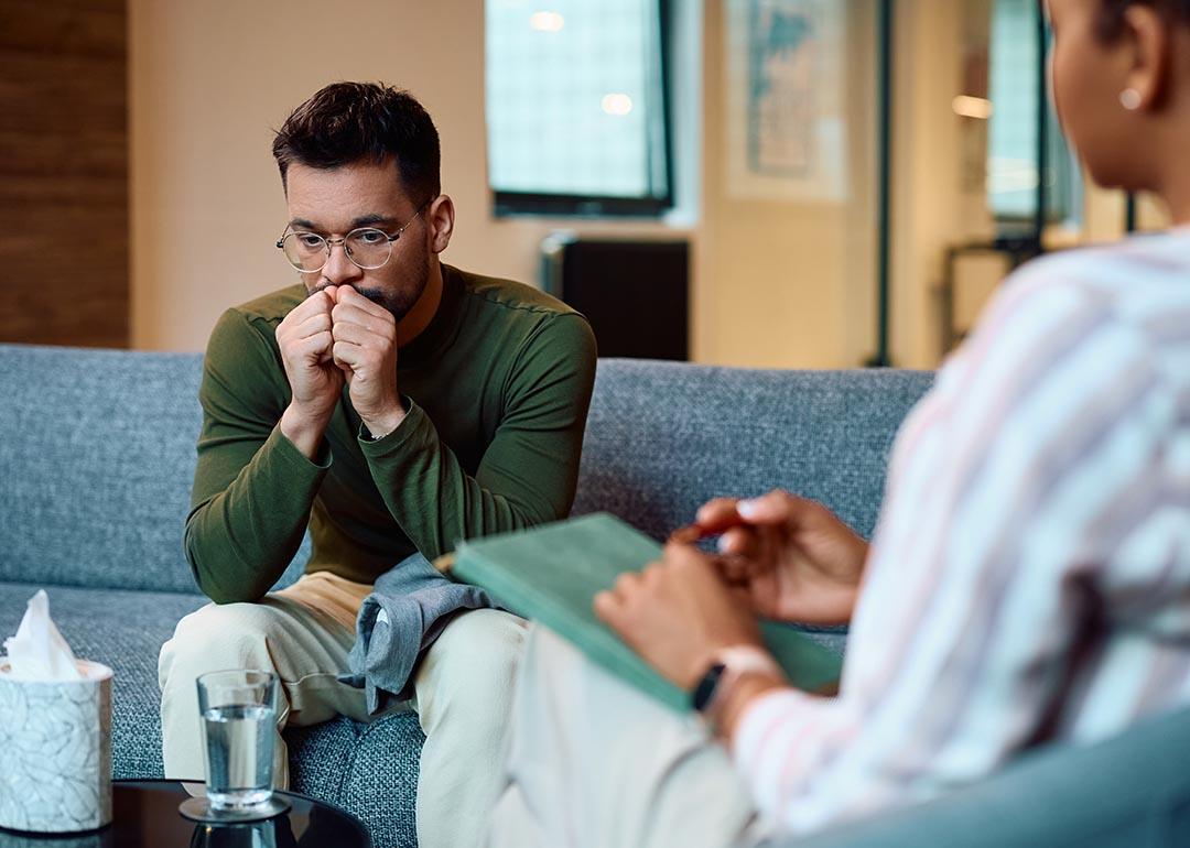 Man in a long sleeve green shirt wearing glasses sits on a blue couch breathing into his hands. A box of tissues is on the table and a therapist's back is in the foreground.