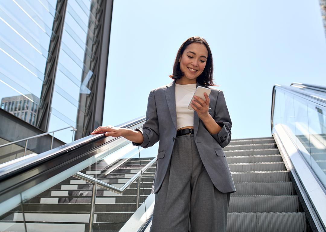 A young female marketing professional smiling at her phone while standing on an escalator.