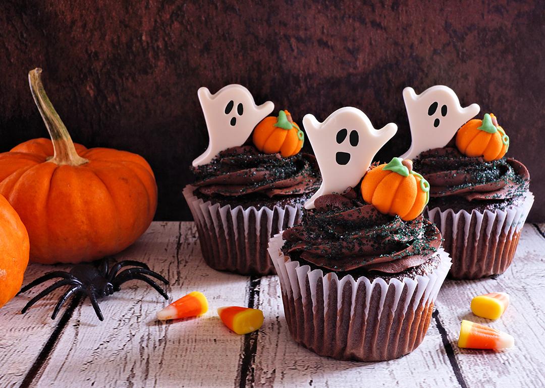 Chocolate cupcakes with ghost toppers beside Halloween pumpkins.