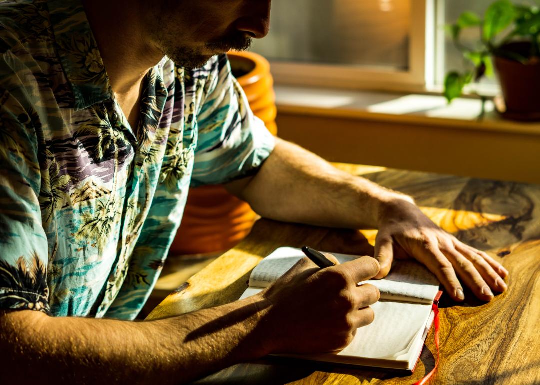 Man wearing a Hawaiian-style shirt sitting next to the window with light coming in, writing in a journal. 