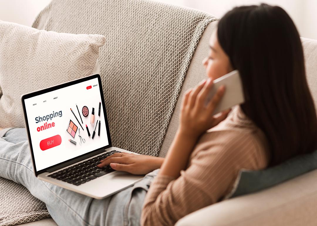 Back view of a woman sitting at home, on the phone, and browsing a shopping website on a laptop.