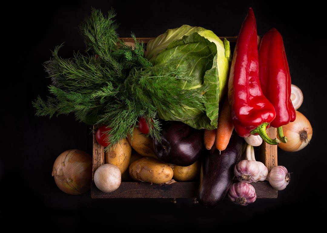 Assortment of beautifully-lit, fresh raw vegetables inlcuding leafy greens and red peppers in a wooden box against black background.