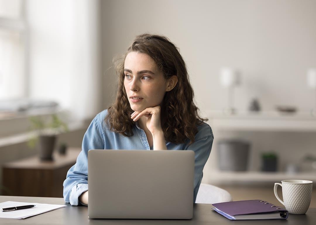 Woman sits at desk with open laptop computer, looking into the distance.