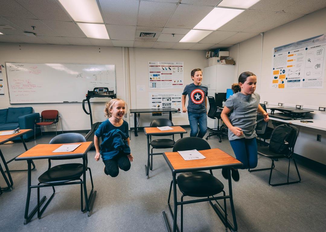 Three elementary-school-age students jumping in the air next to desks in a classroom. 