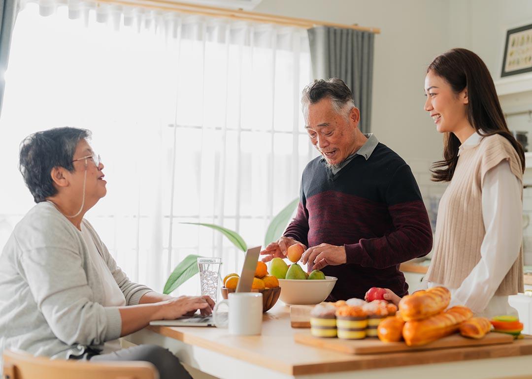 Mother father and adult daughter at the kitchen table talking with window in the background and fruit on the table.