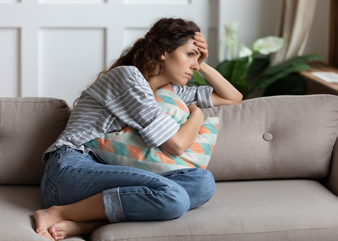 A stressed young woman, sitting at a sofa and cuddling a pillow while lost in thought.