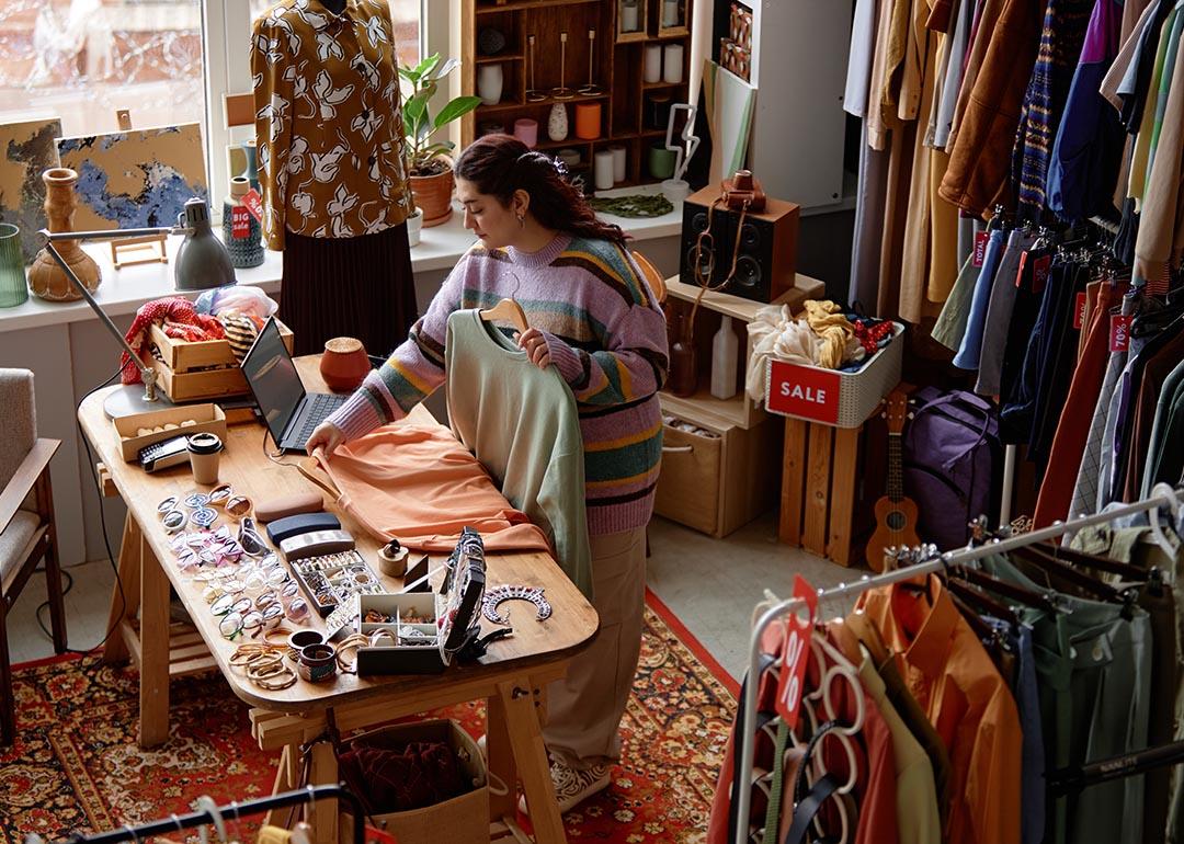 High-angle shot of sales assistant working at checkout desk cluttered with fashion accessories at second hand shop with retro style carpet selling clothes and household goods.