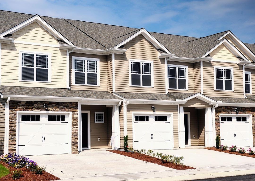 A row of modern townhomes with beige siding, white trim, and stone accents, each featuring a garage, driveway, and small landscaped front yards under a bright blue sky.