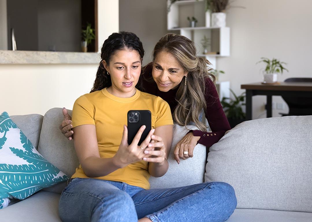 Adolescent daughter on sofa shows interested mum social media on phone screen.