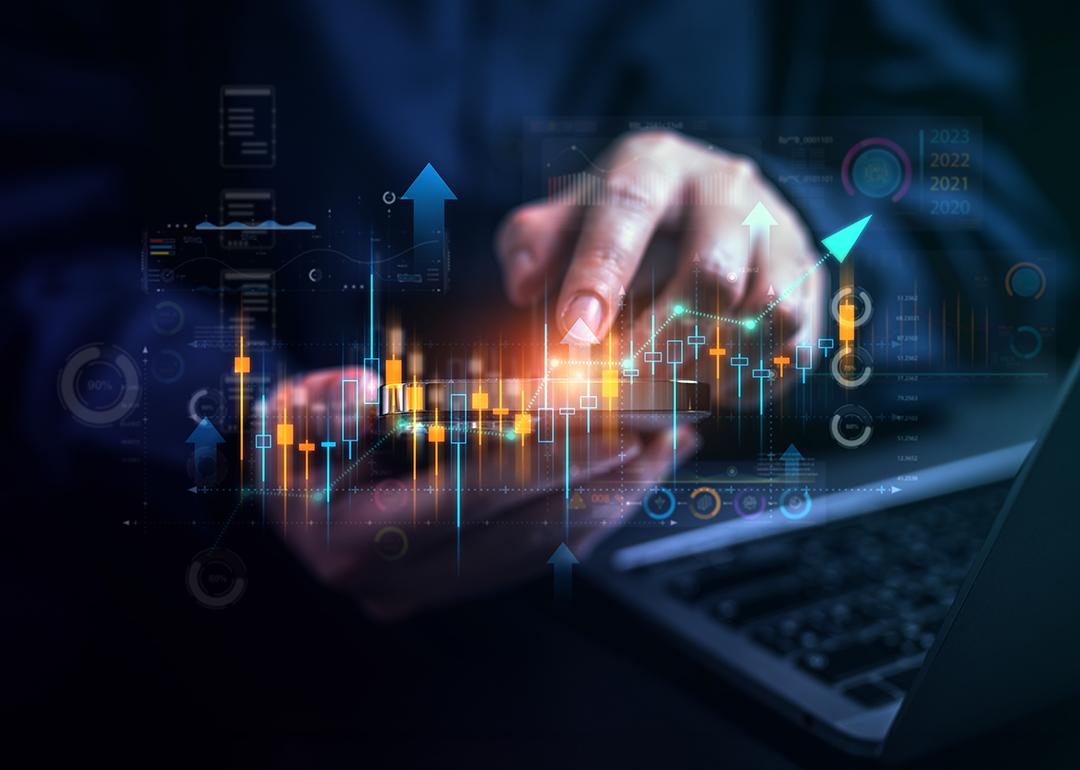 Stock market trading graphics illustrated over a businessperson using a phone and laptop.