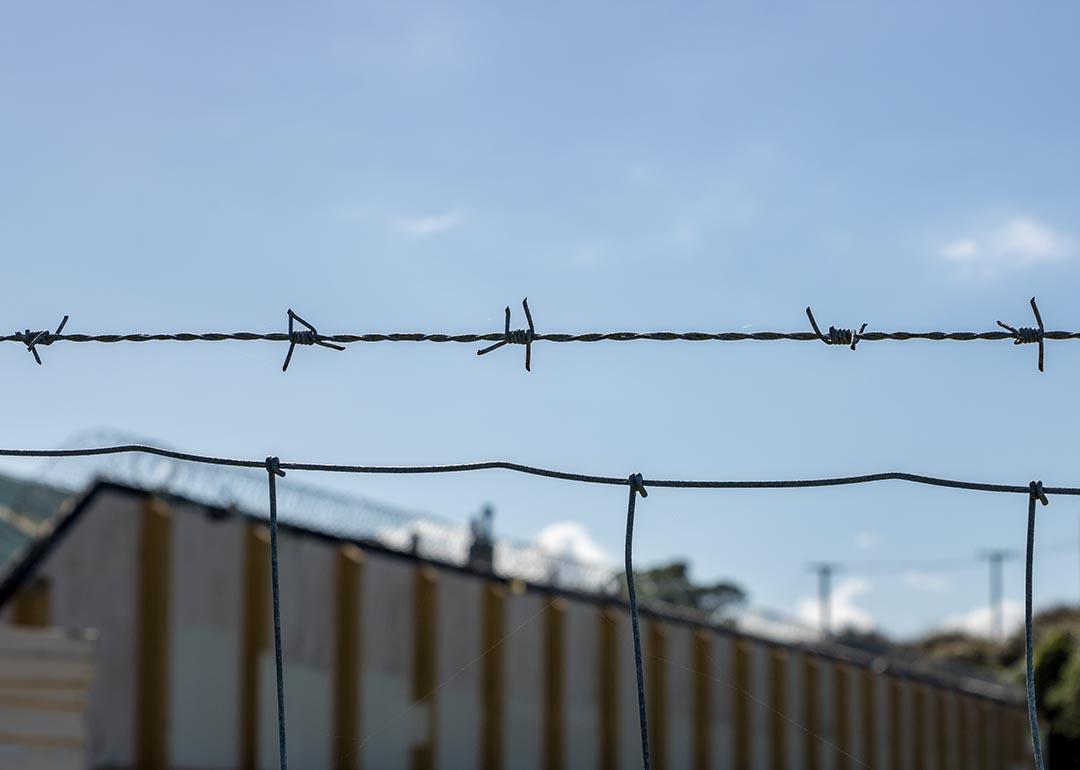 Close up of a single strand of barbed wire above a fence with a correctional facility in the background.