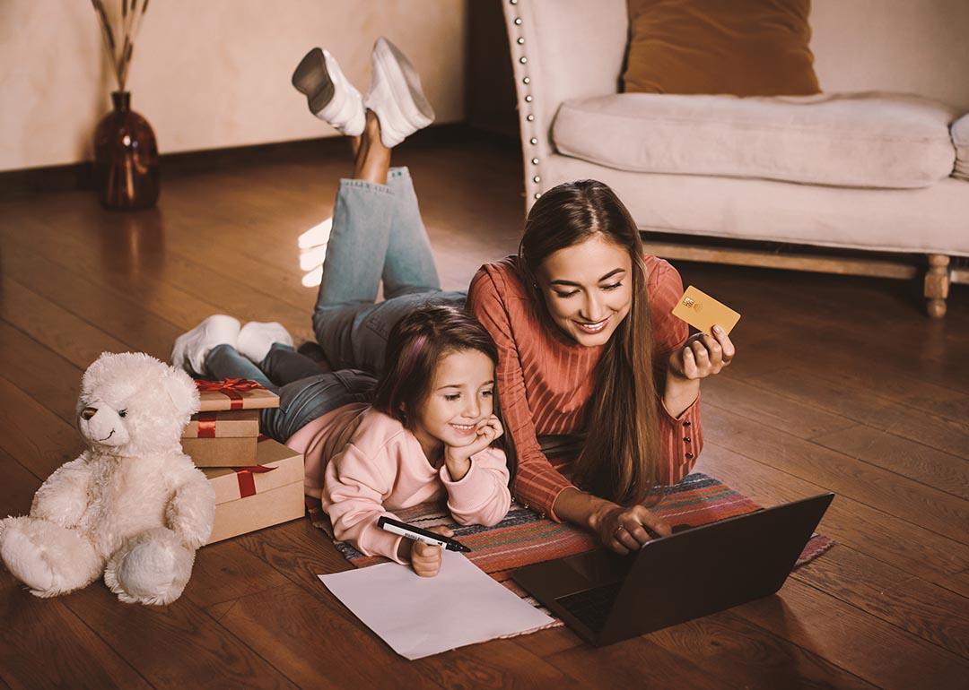 Girl writing wishlist, next to adult woman, both are laying on a rug looking a laptop online shopping while the woman is holding a credit card. There are wrapped gifts next to them along with a teddy bear.