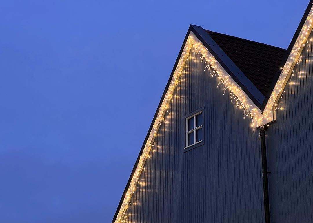 Icicle Christmas Lights hanging along the roof line of a wooden-fronted house. There is a small white window on the house, and a dusky blue sky behind the house.
