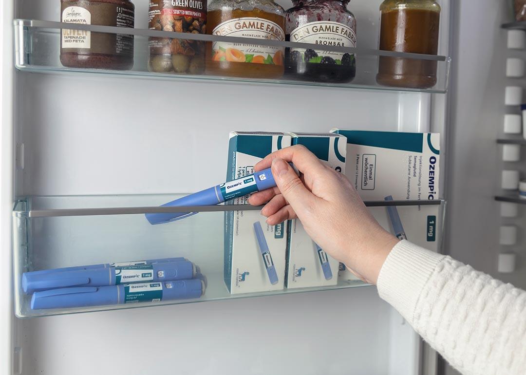 Close up of arm of person wearing white sweater reaching into refrigerator door to pick up an Ozempic injection pen, boxes of Ozempic are stored to the right.