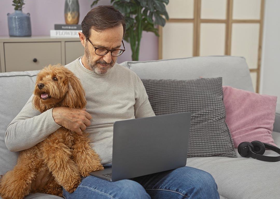 A hispanic middle-aged man cuddles a poodle while using a laptop in a cozy living room setting.