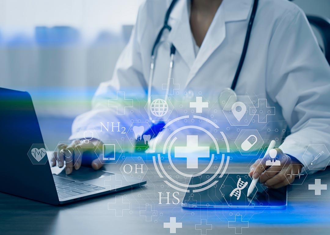 Doctor visible from the neck down sitting at desk with laptop and iPad  working with digital medical interface icons superimposed symbolizing AI. 