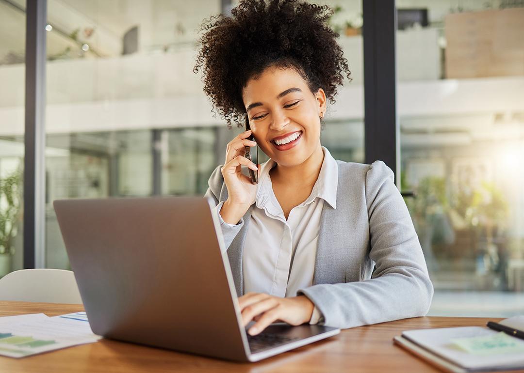 A businesswoman in conversation with someone on her phone and working with a laptop in an office.
