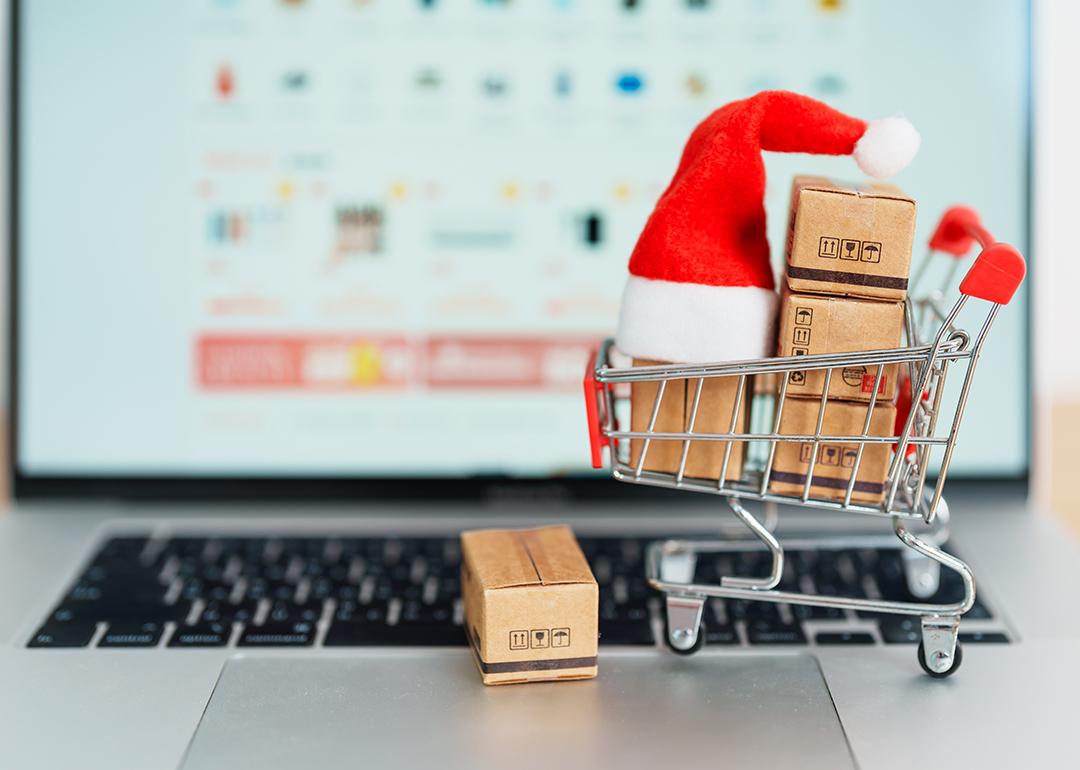 A small shopping cart figure with boxes and a Christmas hat, in front of a laptop displaying a shopping page.