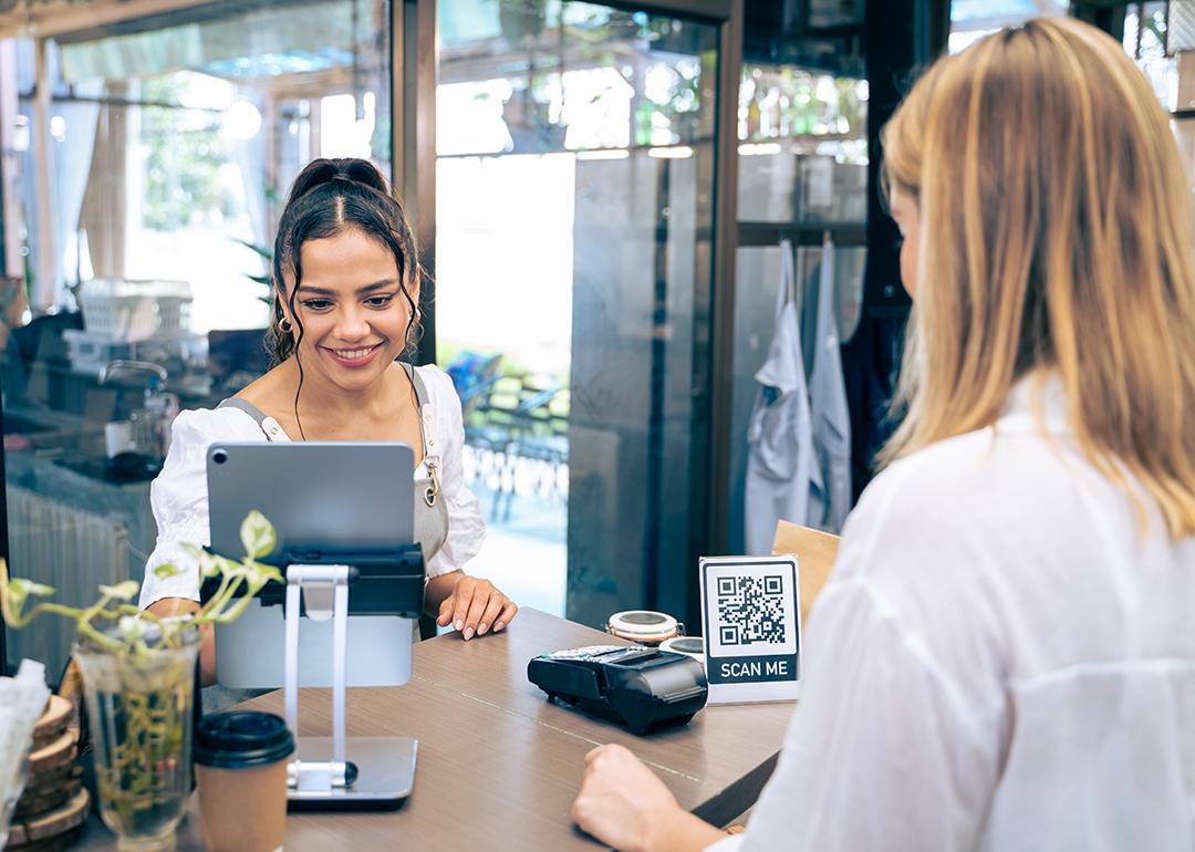 Barista receives a customer lining up to pay in a cafe's counter where a QR code information graphic is displayed.