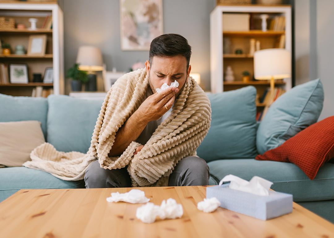 A sick man at home wrapped in blanket sitting on a sofa and blowing his nose.