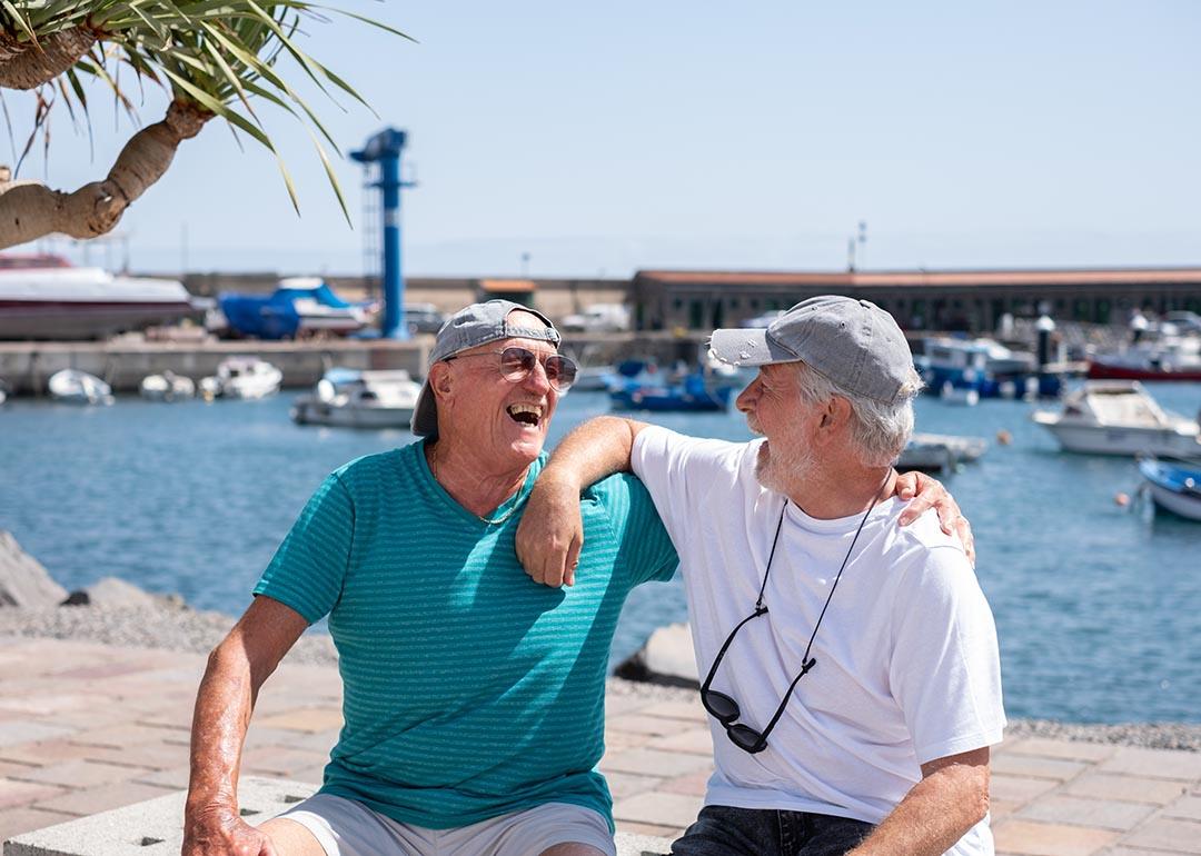 Two senior friends wearing caps and t-shirts in retirement life sitting on a bench at the harbor looking each other laughing happily.
