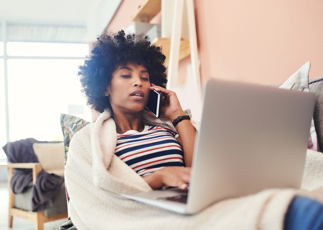 A woman at home on the couch, wrapped in a blanket talking on the phone with a laptop on her lap.