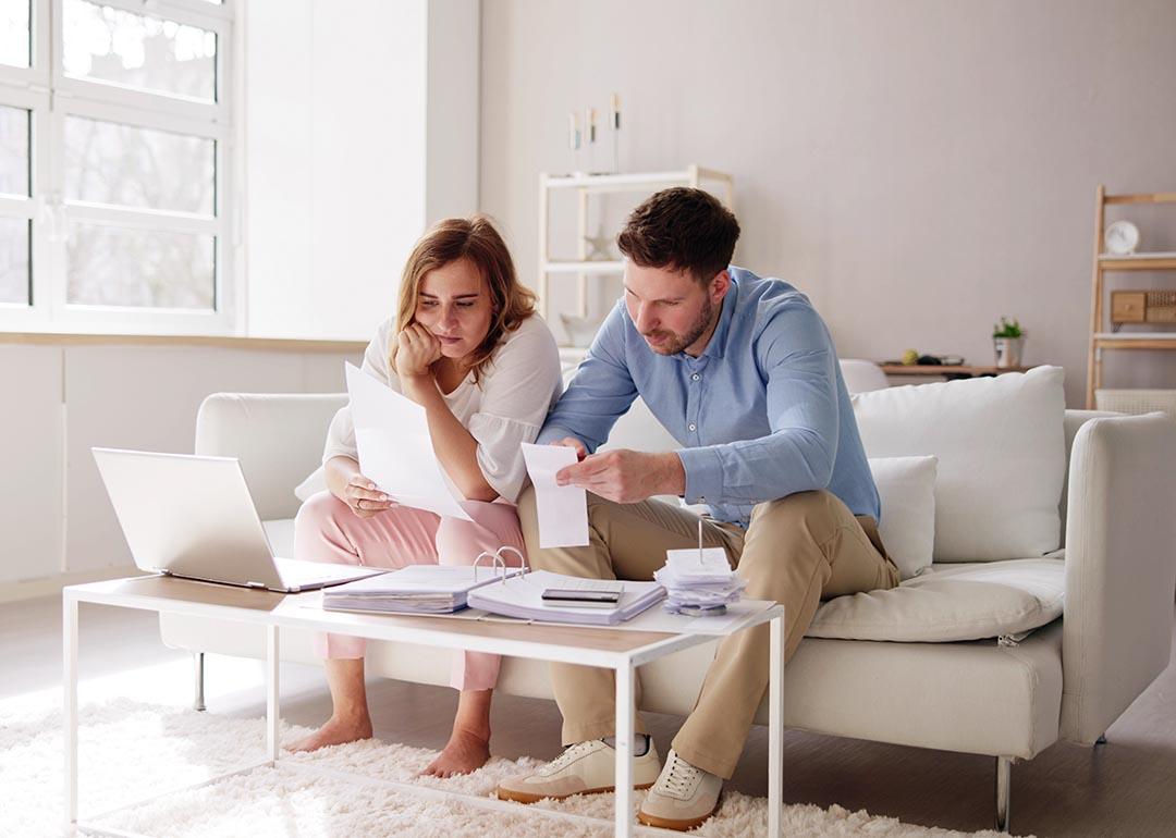 Two people sitting on a white sofa looking at bills and paperwork in a minimalistic living room.