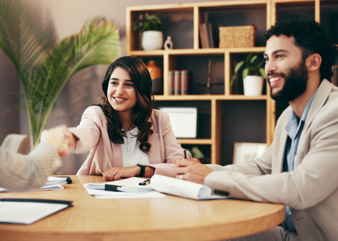 Three business people sit at a table. One smiling person shakes hands with another person, who is mostly out of view. 