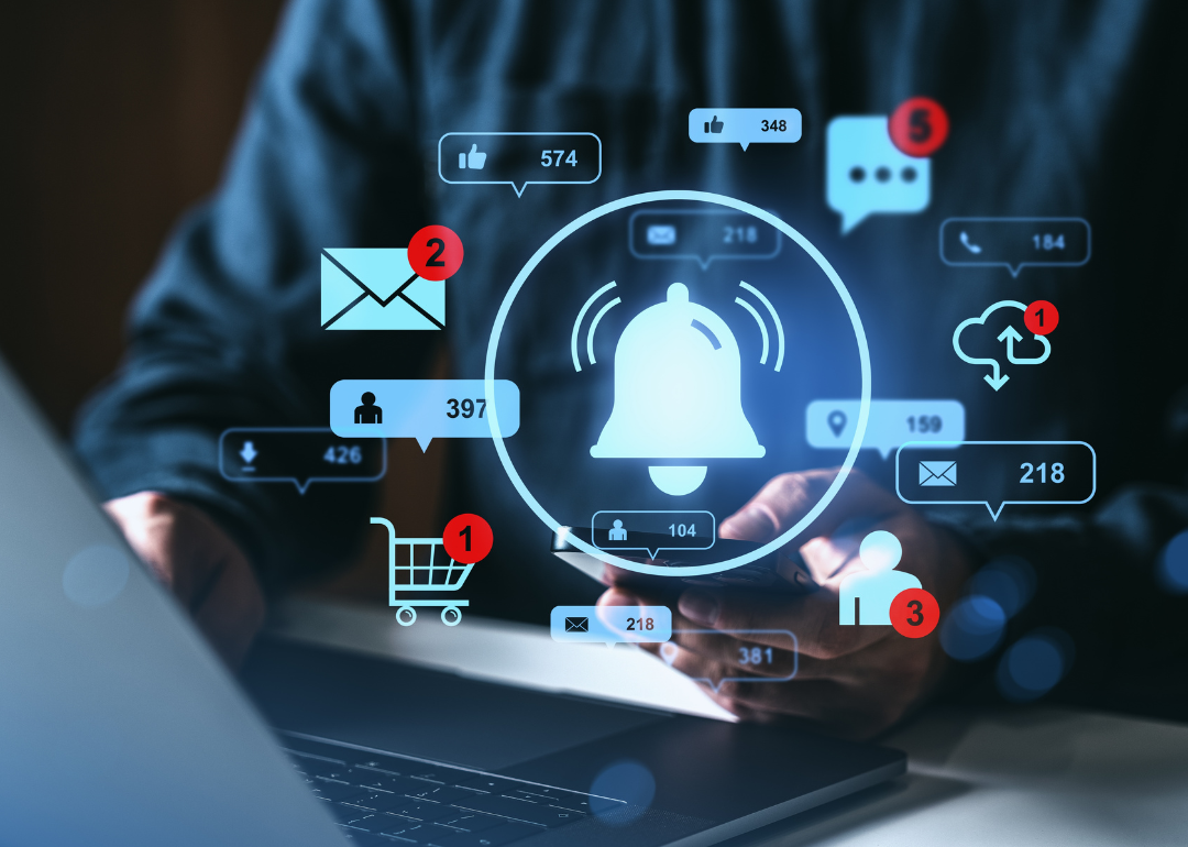 Person using a smartphone in front of a laptop with a various icons, like an alert bell, an email inbox notification, and a shopping cart, overlaying the image. 