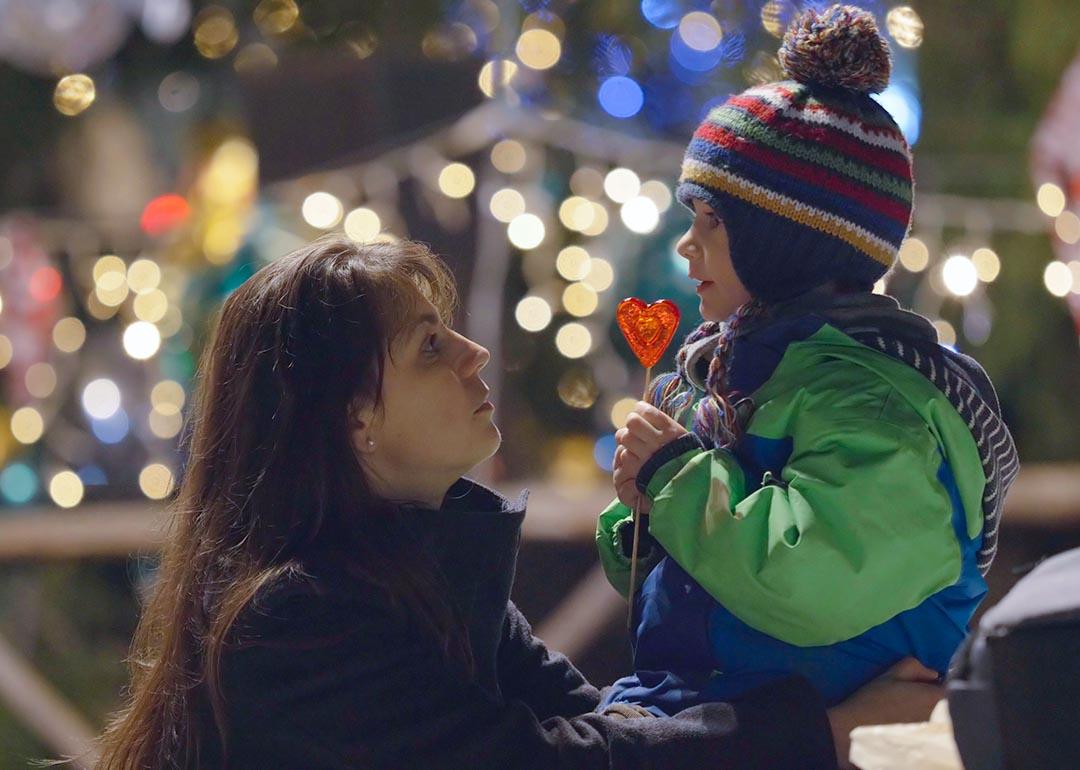 A mother kneels to the height of her child ourdoors and looks into his eyes, he is eating a heart-shaped lollipop and wearing a knitted cap with a pom pom in front of blurry christmas lights in the background.