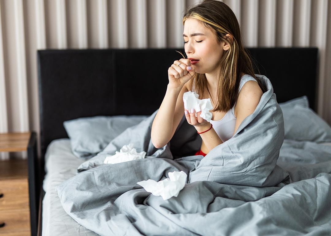 A sick woman wrapped in blanket is sitting on her bed and coughing.