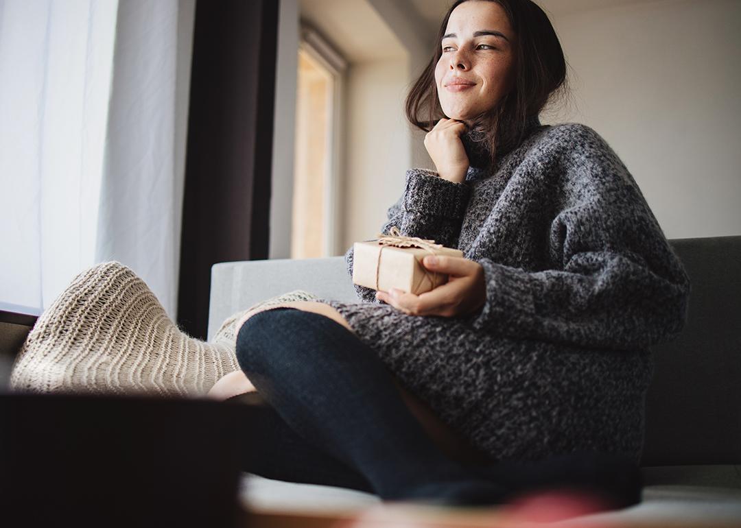 A young woman cozily sitting on a sofa at home holding a small gift box.