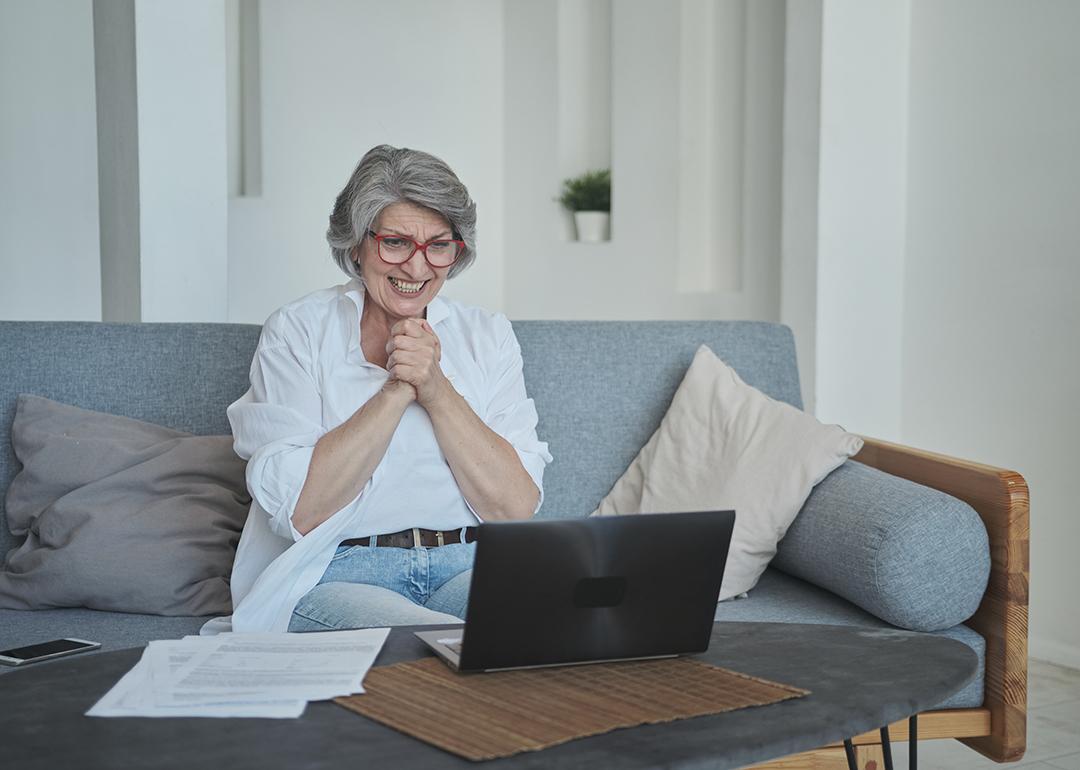 A senior woman sitting on a sofa and excitingly awaits good news on her laptop.