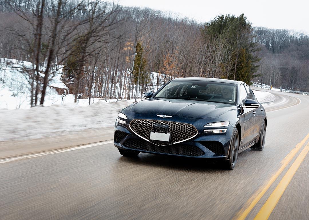 Front view of black Genesis G70 driving on a road in winter with snow and trees in background. 