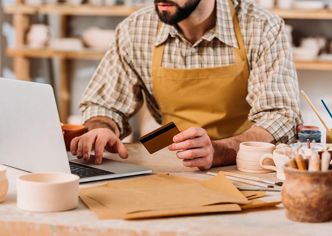 Cropped view of a small pottery business owner holding a credit card and browsing a laptop.