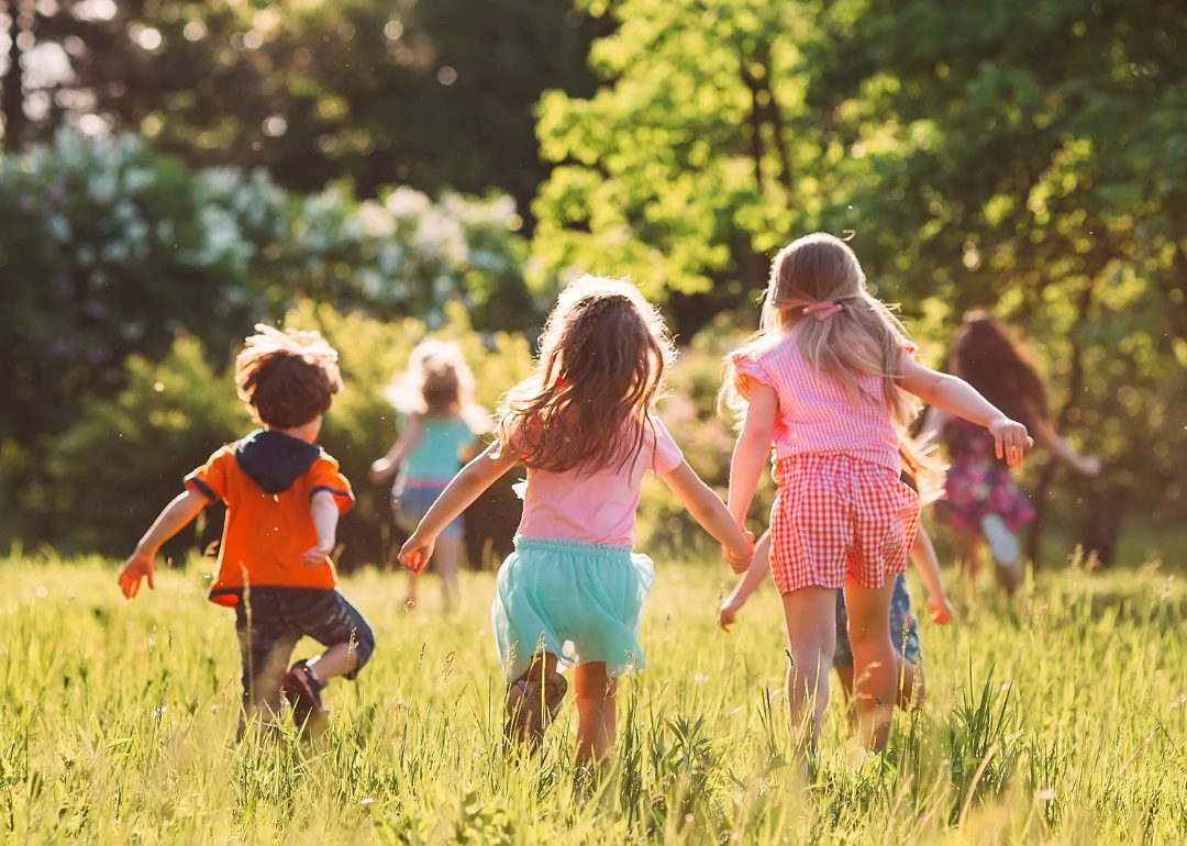 Young children running and playing in a sun-filled meadow with long grass and trees in the distance.