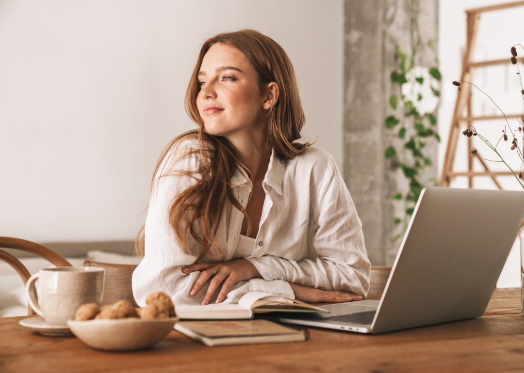 A content-looking woman in her 20s or 30s sits in a sunny room at a table with a laptop, a mug, a notebook, and a bowl of baked goods. 