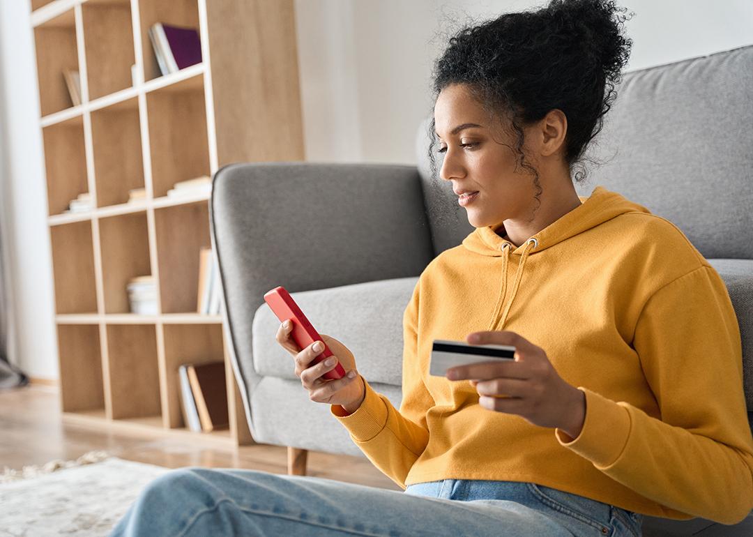 A young woman sitting on the floor of a living room, holding a credit card and browsing her smartphone.