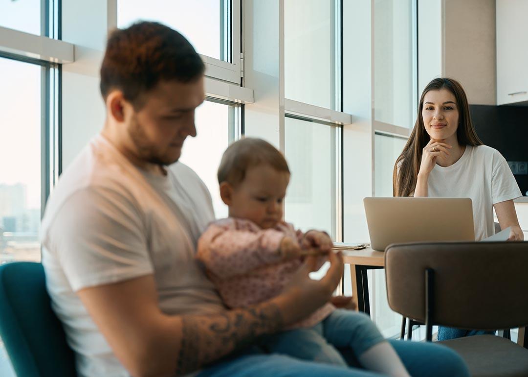 Woman smiling and sitting at table with laptop looking at man holding baby in the foreground.