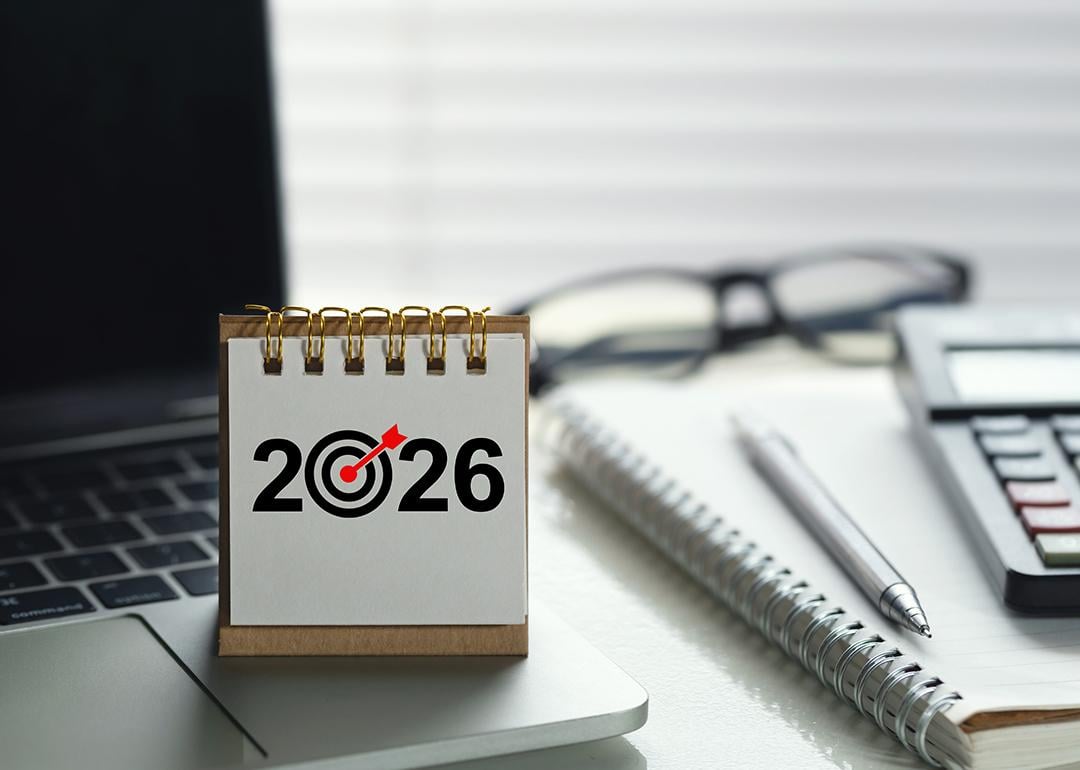 A desk calendar for 2026 with a target icon on top of a laptop among other office desk items.