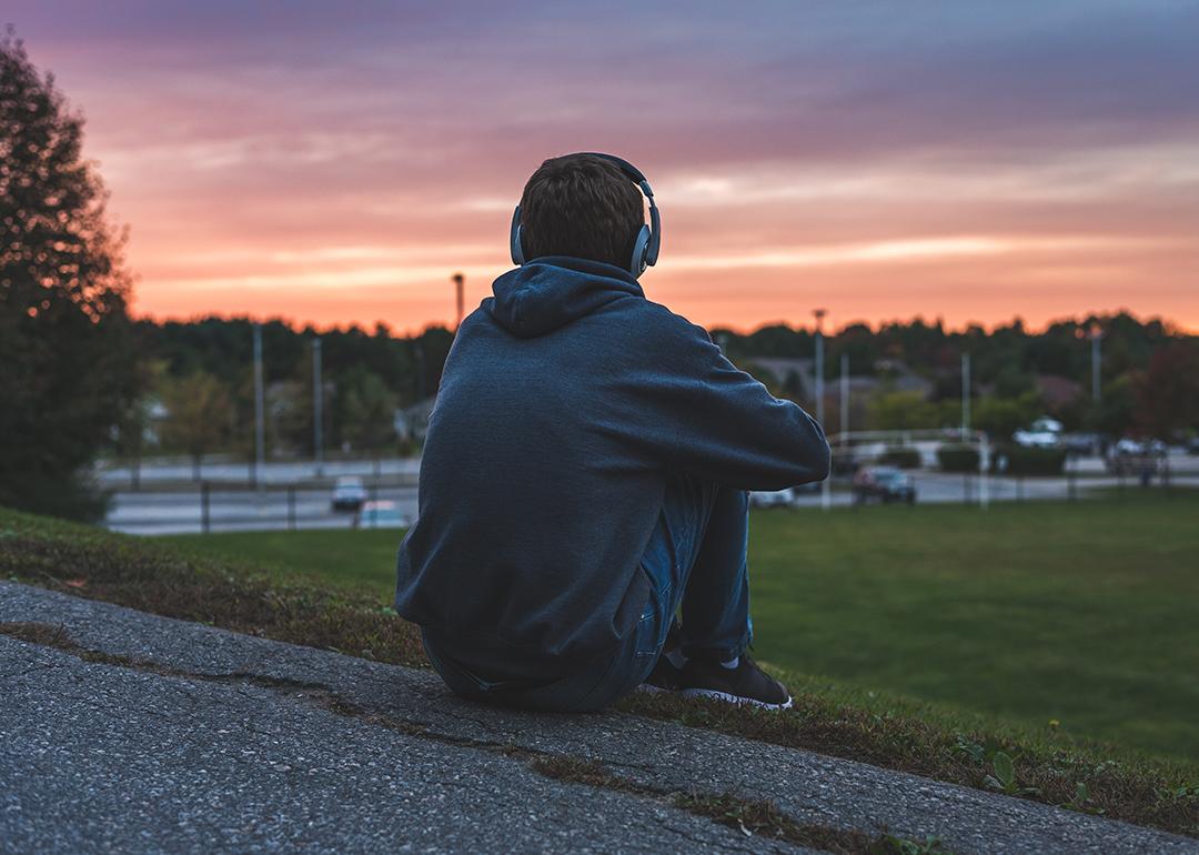 Rear view of a male teenager sitting alone at the top of a hill and watching sunset while listening to headphones.